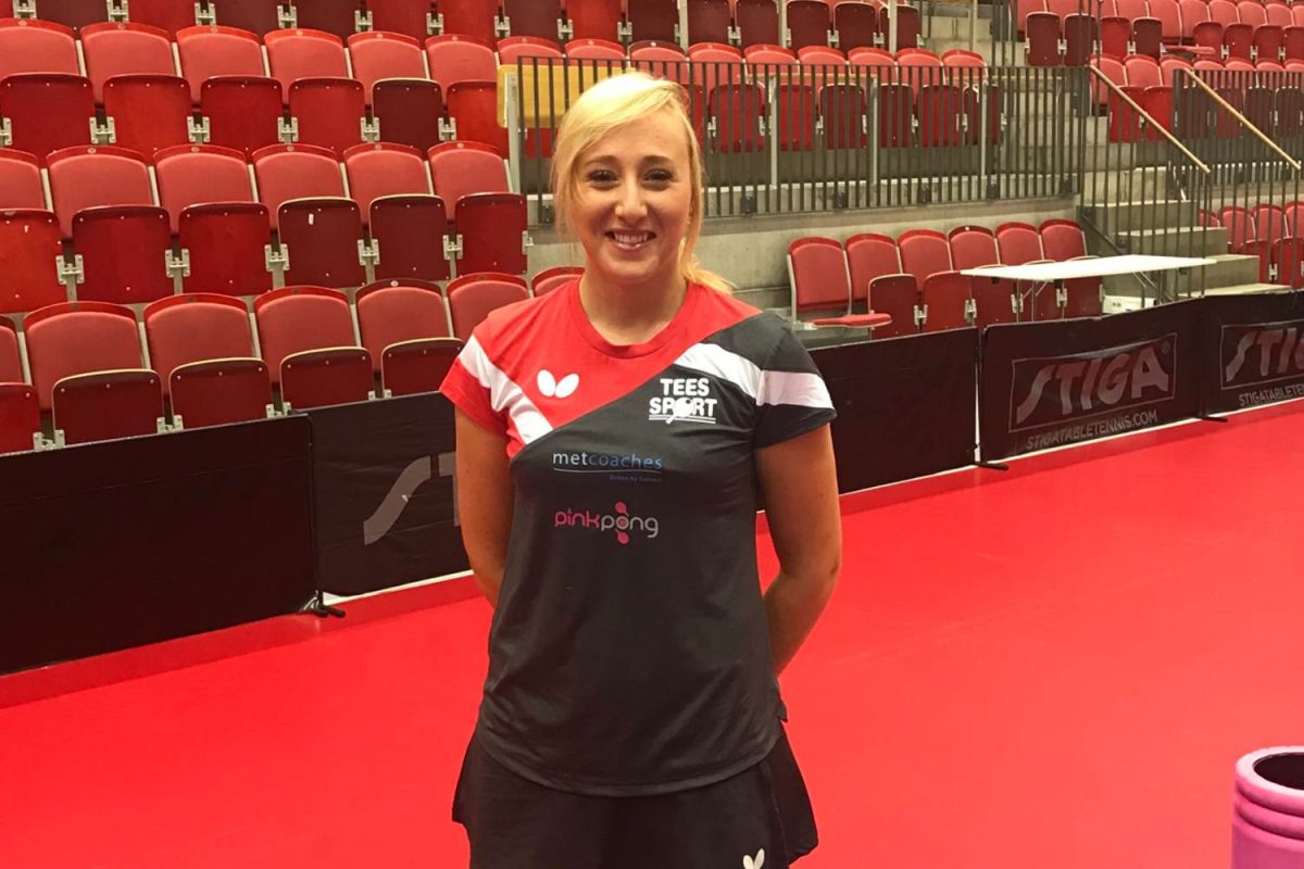 Charlotte Carey standing beside a table tennis table in the red version of her new season kit.